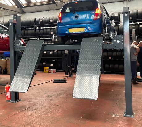 A large four-post car hoist in a workshop, shown with a blue hatchback raised on the right side and a red hatchback on the left. Several people are standing near the right side of the machine, and stacks of tires are visible in the background.